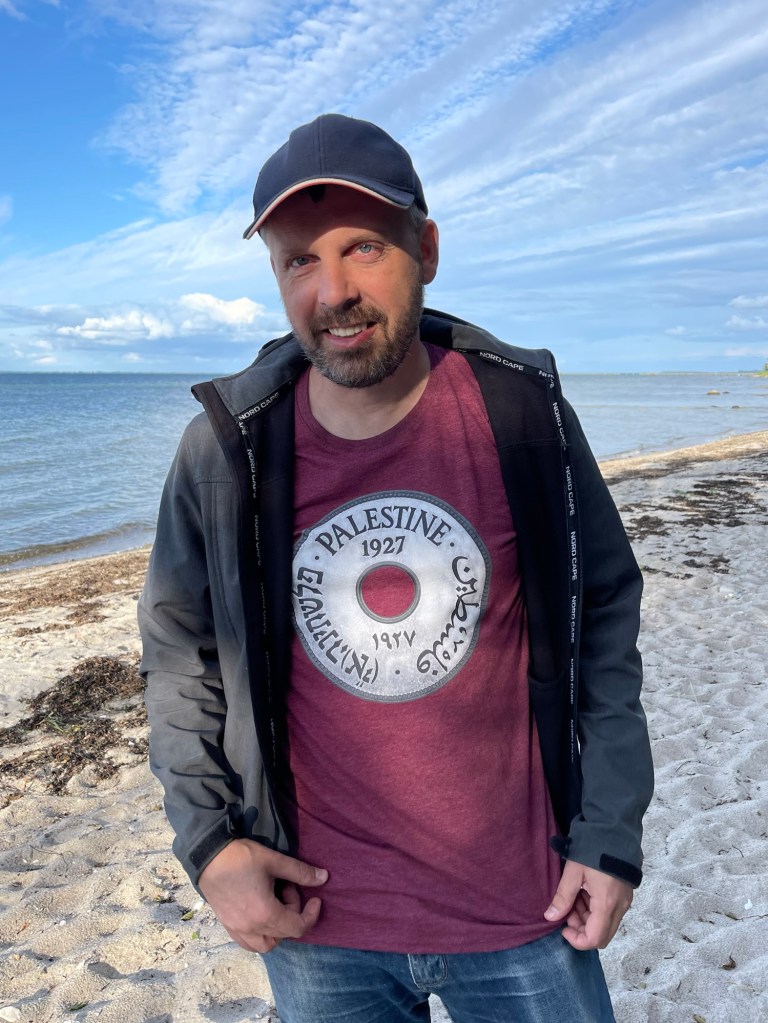 Foto von mir am Strand mit dem Shirt 10-Mils unter einer geöffneten Jacke. Die Münze ist oben mit "Palestine 1927" beschriftet.